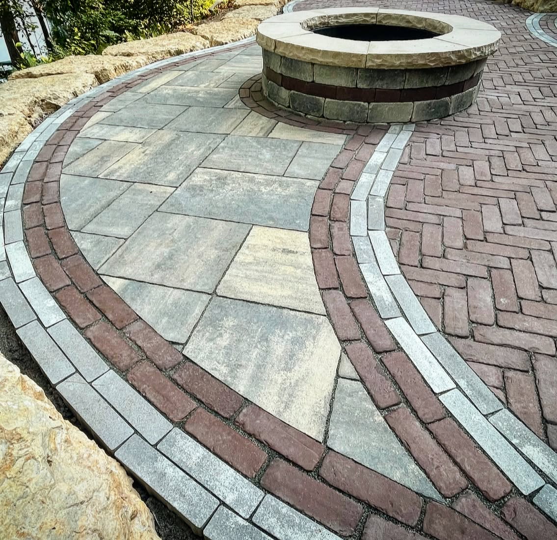 Custom mixed-material paver patio with large-format slabs, herringbone pavers, curved contrasting borders, geometric inlays, and a circular masonry fire pit.