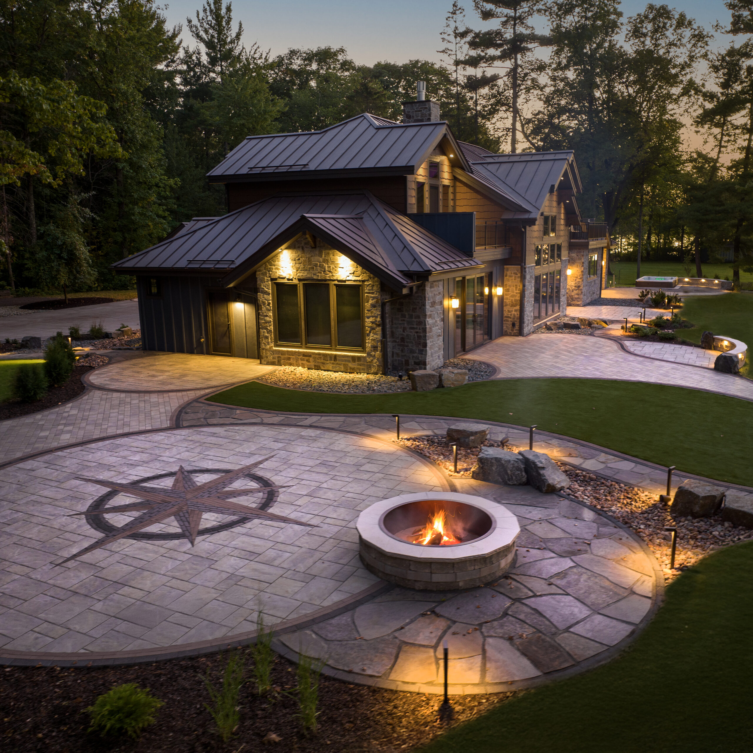 Custom paver patio with a circular fire pit, compass rose inlay, curved walkways, and landscape lighting beside a modern home.