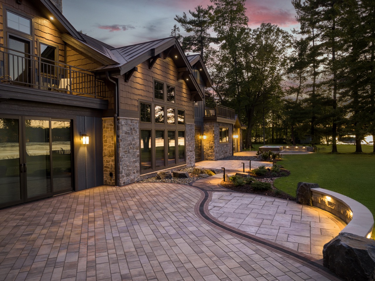 Curved paver patio and walkway with decorative borders, a lighted seating wall, landscaped beds, and lakefront views at sunset.